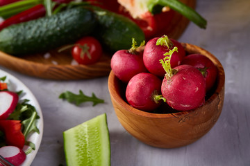 Fresh red radish in wooden bowl in front of plate with vegetables, selective focus.
