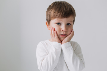 Incredible lovely little boy with big light eyes dressed white shirt holds hands on the cheeks and looking at camera with surprised emotions, isolated background