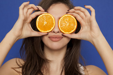 Smiing young woman posing with slices of oranges on her face on blue background