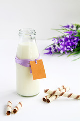 milk bottle with tag for writing wishes, next to her chocolate wafer tubes on white background and right side lilac flowers