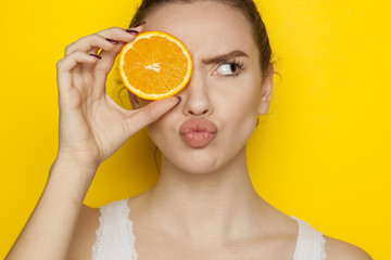 Young woman posing with slice of orange on her face on yellow background
