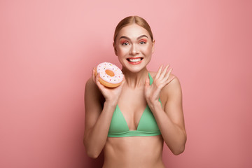 Waist up portrait of rejoicing woman in bathing costume holding tasty donut. Isolated on rose background