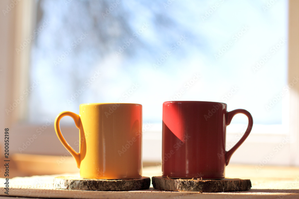 Wall mural sunny atmosphere of spring morning/ two mugs of a hot drink on table in the background of window with silhouette of tree and blue sky