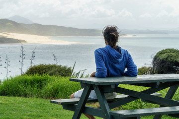 Fototapeta premium rear view of man sitting on bench and looking at ocean, Omapere, New Zealand