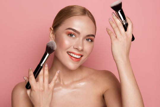Portrait of attractive woman holding blush brushes. She is looking at camera with delight. Isolated on rose background