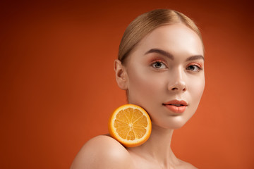 Portrait of attractive woman with pure derma holding half of juicy orange on her shoulder. She is looking at camera with serenity. Copy space in left side. Isolated on background