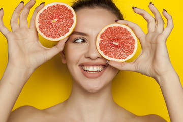 Happy young woman posing with slice of red grapefruit on her face on blue background