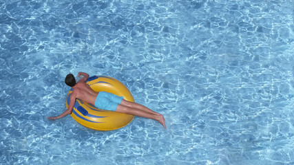 Young man is floating on yellow inflatable air ring/circle in pool with blue water, vacation concept with space for text. Top view.