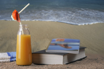 orange smoothie, book, magazine and towel on hot summer sand . Vacation concept