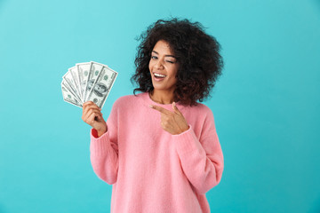 Portrait of american successful woman 20s with afro hairstyle holding lots of money dollar ﻿banknotes, isolated over blue background