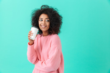 Portrait of american woman 20s with afro hairdo looking aside while drinking takeaway coffee or tea from paper cup, isolated over blue background