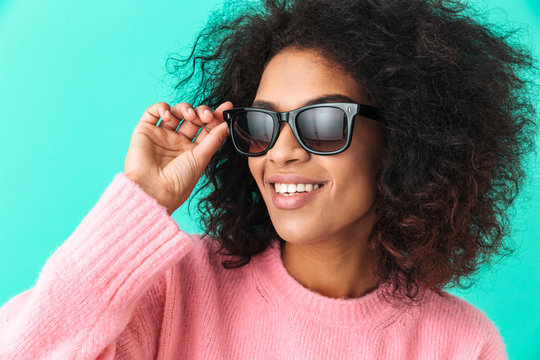 Multicolor image of american woman 20s with afro hairstyle smiling and wearing black modern sunglasses, isolated over blue background