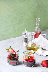 Beetroot and strawberry salad served with arugula and nuts in glass jars with cloth, pepper and bottles of fruit ocet and olive oil over grey table with green wall as background. Healthy eating