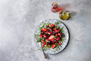 Beetroot and strawberry salad served with arugula and nuts on ceramic plate with bottles of fruit ocet and olive oil over grey texture background. Top view, copy space. Healthy eating - obrazy, fototapety, plakaty