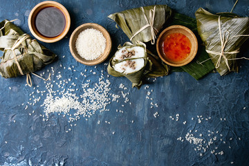 Asian rice piramidal steamed dumplings from rice tapioca flour with meat filling in banana leaves. Ingredients and sauces above over blue texture background. Top view, space.
