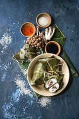 Asian rice piramidal steamed dumplings from rice tapioca flour with meat filling in banana leaves served in ceramic bowl. Ingredients and sauces above over blue texture background. Top view, space.