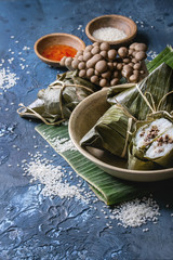 Asian rice piramidal steamed dumplings from rice tapioca flour with meat filling in banana leaves served in ceramic bowl. Ingredients and sauces above over blue texture background. Close up, space.