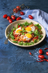 Traditional Israeli Cuisine dishes Shakshuka. Fried egg with vegetables tomatoes and paprika in ceramic plate with cloth, herbs and ingredients above over blue texture background. Copy space.
