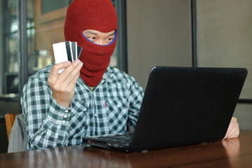 Hands of masked hacker wearing a balaclava holding credit card between stealing data from laptop. Internet crime concept.