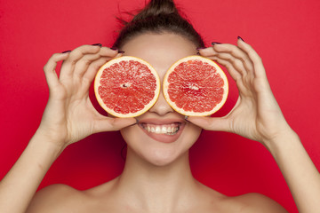 Happy young woman posing with slices of red grapefruit on her face on red background