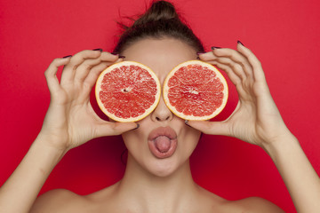 Happy sexy woman posing with slices of red grapefruit on her face on red background