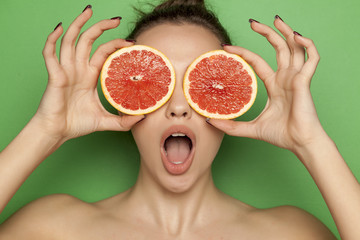 Surprised young woman posing with slices of red grapefruit on her face on green background