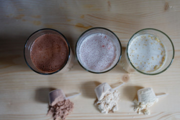 Three milk whey protein shakes in glass cups are arranged in a row with three scoops of protein on a wooden table. Top view three fresh milk, chocolate, blueberry and banana drinks near plastic spoons