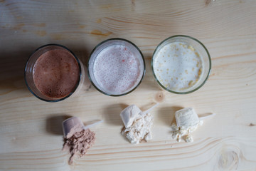 Three milk whey protein shakes in glass cups are arranged in a row with three scoops of protein on a wooden table. Top view three fresh milk, chocolate, blueberry and banana drinks near plastic spoons