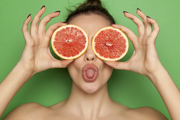 Sexy young woman posing with slices of red grapefruit on her face on green background