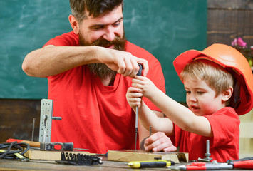 Boy, child busy in protective helmet learning to use screwdriver with dad. Father, parent with beard teaching little son to use tool screwdriver. Teamwork and assistance concept.