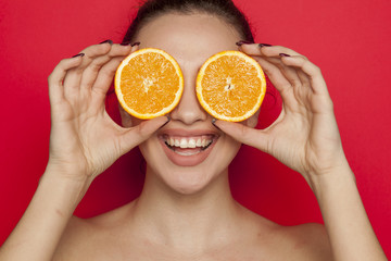 Happy young woman posing with slices of oranges on her face on red background