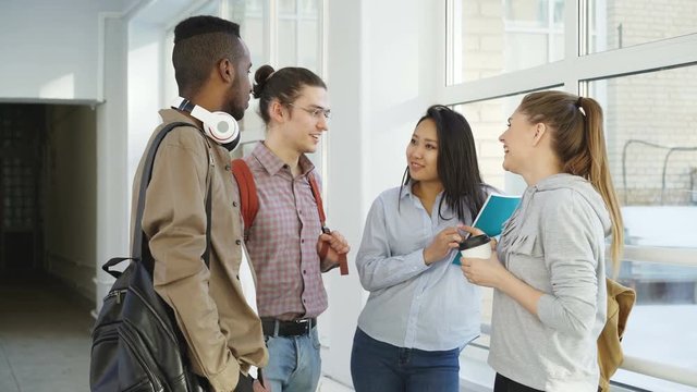 Multi-ethnic group of four students standing in wide lighty glassy hall in college talking to each other in positive way smiling ant laughing dinking coffee