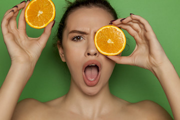 Surprised young woman posing with slices of oranges on her face on green background