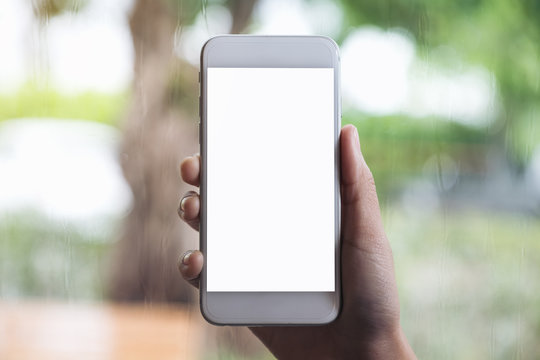 Mockup image of a hand holding white mobile phone with blank desktop screen with blur raining background