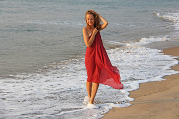 A beautiful girl with blond hair, in a red dress, walks along the seashore and smiles. Summer girl...