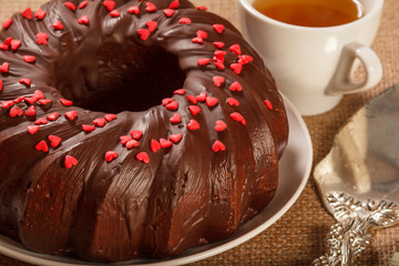 Homemade chocolate cake decorated with small hearts made of caramel on plate, silver cake lifter beside it and cup of tea