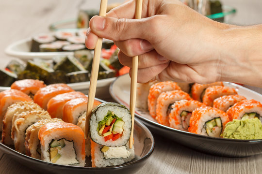 Male hand with two  chopsticks holding Hosomaki roll with vegetables and different  sushi rolls with seafood on ceramic plates on the background