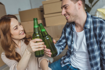 young couple clinking bottles of beer at new apartment, relocation concept