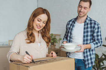 smiling woman signing cardboard box with husband with dishes near by, moving home concept