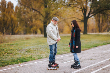 Fototapeta premium A happy pensioner stands on roller skates on the road with his daughter and enjoys good weather in the autumn park.
