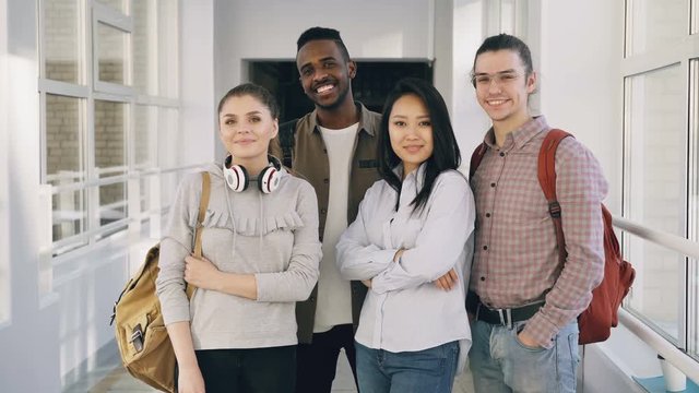 Portrait of four smiling positive attractive multi-ethnic male and female students standing in spacious white corridor in university looking at camera
