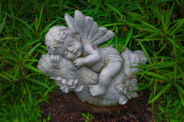 LITTLE ANGEL SLEEPS.  Sculpture of a little angel sleeping on tree a branch. It is placed on top of an old tree stump surrounded with green leaves.