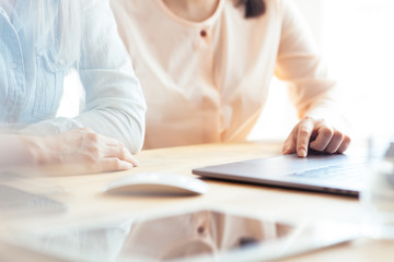 Close up shot of two female businesswomen working with hands on a laptop keyboard in an office in sunlight at a wooden table
