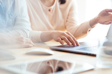 Close up shot of two female businesswomen working with hands on a laptop keyboard in an office in sunlight at a wooden table