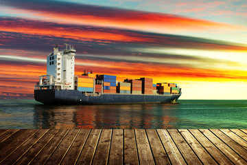 View from wooden pier with container ship on sea during sunset.