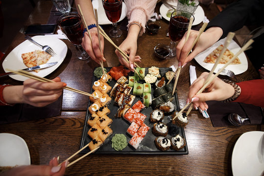 A set of sushi rolls on a table in a restaurant. A party of friends eating sushi rolls using bamboo sticks.