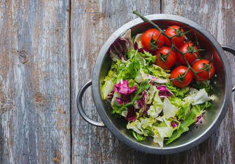 Fresh salad in the steel aluminium colander. Lettuce, arugula and red cherry tomatoes on the vine. Vegan summer food in the kitchen on old vintage wooden background. Vegetarian and healthy vegetables.