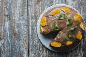 Homemade sponge cake with chocolate cacao ganache frosting. Slices of kiwi and orange on the dessert. Baked pastry in the kitchen on the old vintage wooden background.