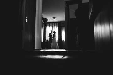 Beautiful wedding couple is hugging near the window. Bride and groom silhouettes on the dark red background. Romantic meeting in the wedding morning. Black and white.