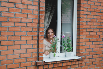 Fototapeta na wymiar Beautiful bride is looking out the window waiting her groom. Young blonde girl in white wedding dress and elegant hair accessories. Wedding portrait from outside. Red bricks house and flowers plants.